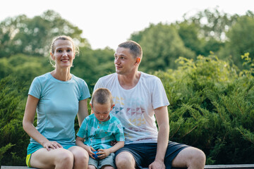 Parents and twins boy and girl sitting on wooden bridge in a park. Sunny day, good mood. Happy joyful young family father, mother and little son having fun outdoors, playing together in summer.
