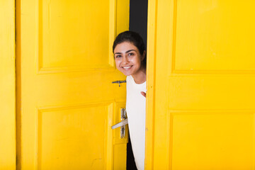 Young smiling indian woman peeking through yellow open door