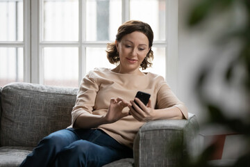 Smiling older Caucasian woman sit rest on couch at home text or message online on modern cellphone gadget. Happy mature female relax browse internet on smartphone device. Elderly technology concept.