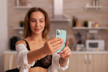 Fototapeta na wymiar Portrait of beautiful woman looking at phone camera while taking selfie in home kitchen weraring sexy underwear. Seductive woman with tattoos using smartphone wearing temping lingerie in the morning.
