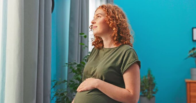 A woman with beautiful red hair is standing by the curtains, looking ahead, holding her pregnant belly, imagining her baby, thinking ahead, thinking of a name for the child