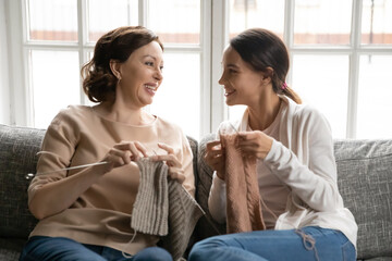 Smiling old Caucasian mother and adult daughter sit relax on couch at home have fun rest knit with...