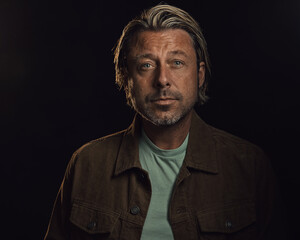 Middle-aged man with a stubble and blond hair wears a brown corduroy jacket with a light green t-shirt.