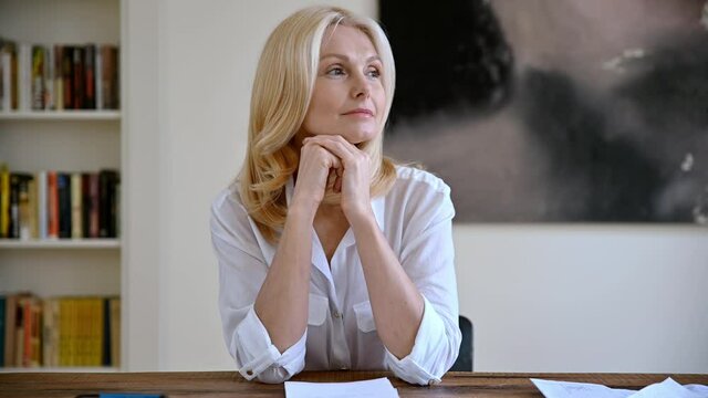 Video portrait of a positive elegant successful mature blonde woman in white stylish shirt, sits at the desk at office, taking a break from work, looking to the side then et camera, smiling friendly
