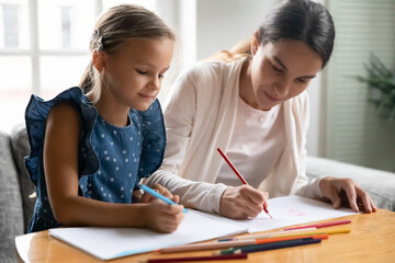 Caring young Caucasian mother and little teen daughter sit at table at home draw paint in album with pencils. Loving happy mom have fun enjoy learning hobby activity with small girl child together.