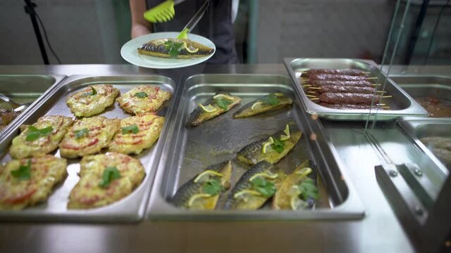 In a school cafeteria or buffet on the food service line, black-gloved attendants place baked fish with lemon and mackerel on a plate.