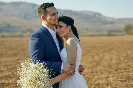 A handsome groom embracing his bride and smiling at the field with great view of hills. Outdoors. Nature