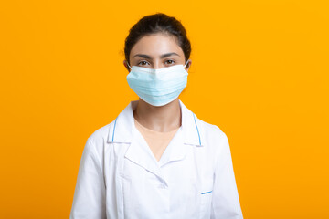 Portrait of indian doctor woman wearing white medical gown and face mask isolated on yellow background.