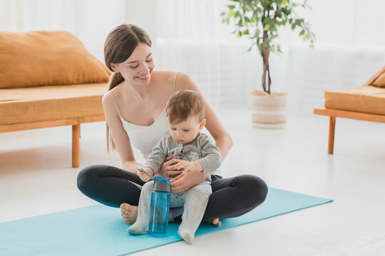 Young Caucasian Mother Doing Fitness Exercises After Labor And Baby Delivery On Mat, Meditating With Her Little Toddler Son Infant Baby At Home On Lockdown. Shaping Body In Postnatal Period