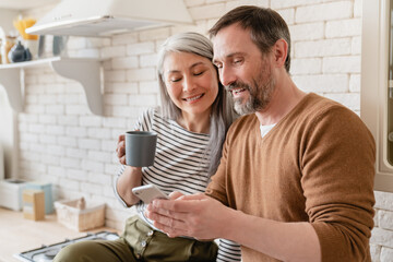 Mature family middle-aged wife and husband couple using smart phone watching videos, social media, reading news on cellphone at home in the kitchen together. - obrazy, fototapety, plakaty