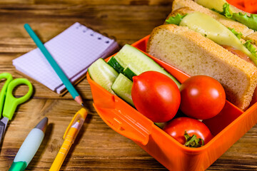 Different stationeries and lunch box with sandwiches, cucumbers and tomatoes on wooden table