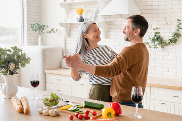 Cheerful mature middle-aged couple family wife and husband cooking romantic dinner for date, drinking wine and dancing together in the kitchen,preparing vegetable salad. Togetherness and relationship