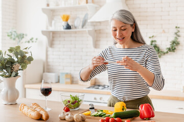 Happy mature middle-aged woman wife mother blogger taking photo of her cooking process while preparing vegetable salad vegetarian food meal in the kitchen at home. Food blogging