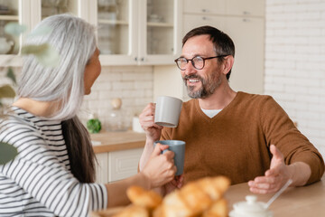Happy middle-aged mature couple family wife and husband having breakfast brunch together in the kitchen at home drinking hot beverages. Love and relationship concept