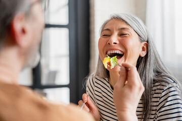 Caring mature husband feeding his middle-aged wife with a vegetable salad in the morning for breakfast on romantic date dinner. Happy couple family eating together vegetarian food