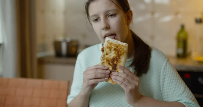 View of teen age girl eating her tasty pita or shawarma. Pretty girl smiles to camera. Beautiful domestic kitchen background. A day in a life