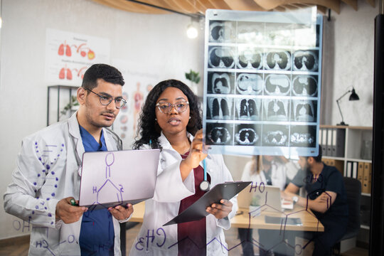 Group of two multiracial doctors, standing behind glass wall, using laptop while examining tomography of patient during consilium. International colleaguesdiscussing various way of treatment. - Powered by Adobe