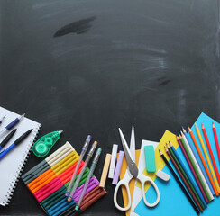 Top view of school items: notebook, pens, plasticine, crayons, erasers, scissors, pencils on the blackboard. Back to school, flat lay with copy space 