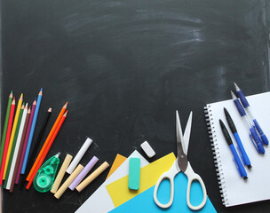 Top view of school items: notebook, pens, plasticine, crayons, erasers, scissors, pencils on the blackboard. Back to school, flat lay with copy space 