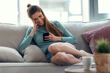 Obraz premium Pretty young smiling woman using mobile phone while taking a break sitting on a couch at home.
