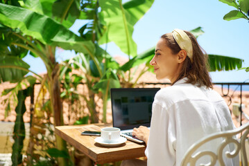 Young beautiful mixed race woman sitting on terrace at morning in hotel, using laptop computer while enjoying vacation