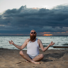 Fototapeta premium A bald man with a red beard practices yoga on the beach at sunset. A funny dude in a T-shirt and sunglasses meditates on the seashore.