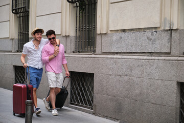 Gay couple carrying suitcases and going to the hotel embraced in a city street.