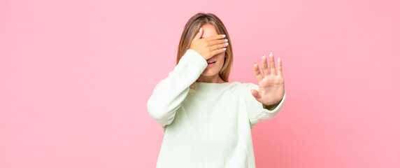 blonde pretty woman covering face with hand and putting other hand up front to stop camera,...