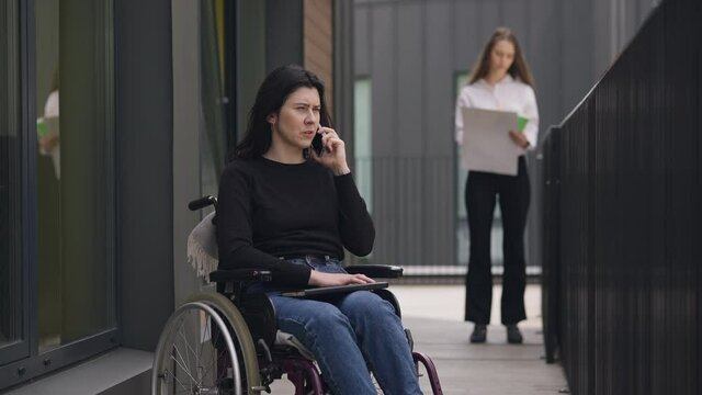 Portrait of confident disabled young woman in wheelchair talking on the phone with blurred colleague analyzing paperwork at background. Successful Caucasian coworkers outdoors. Disability and business