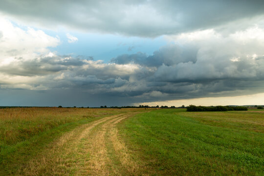 Rural road through meadow and rainy cloud on sky