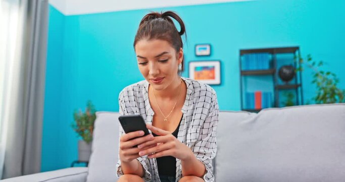 Young woman sits on sofa in living room holding phone in hands, girl looks at pictures on smartphone, smiles, chats with friends, surfs the internet, spends her free time in front of the screen