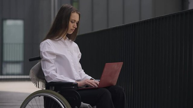Young professional beautiful disabled woman sitting in wheelchair messaging online on laptop. Portrait of confident Caucasian female employee typing on keyboard thinking planning business idea