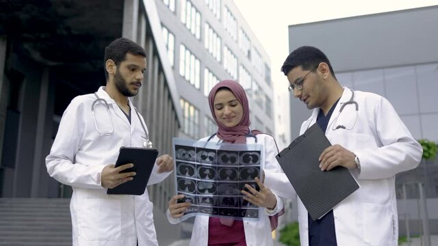 Team of professional three male and female Arabian doctors examining patient's tomography scans while standing outside the modern hospital building. Doctors discussing x-ray image