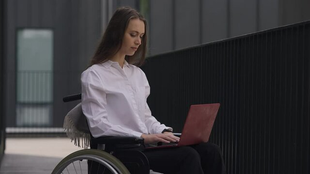 Brunette disabled confident woman messaging online on laptop looking at camera and smiling. Portrait of charming paralyzed Caucasian manager in wheelchair posing outdoors in slow motion