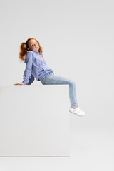 One cute red-headed girl in casual clothes sitting on big box isolated on white studio background. Happy childhood concept. Sunny child. Looks happy, delighted