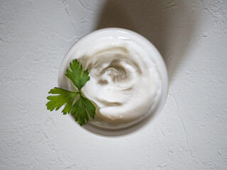 Close-up of fresh sour cream in white sauce bowl. Whirlpool of sour cream. Parsley leaves for decoration. Light concrete background. Delicious and natural dairy product. Flat lay