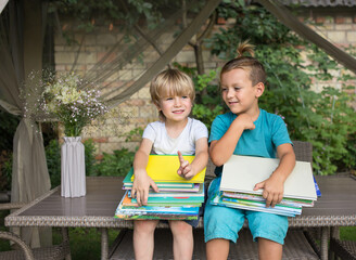 two inquisitive preschoolers are sitting on table in yard, holding stacks of children's books on...