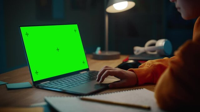 Smart Young Boy Researching Homework on Laptop Computer with Green Screen Display. Teenager Browsing Educational Research, Writing in Notebook, Studying School Material.
