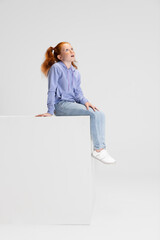 One cute red-headed girl in casual clothes sitting on big box isolated on white studio background....