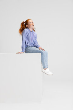 One cute red-headed girl in casual clothes sitting on big box isolated on white studio background. Happy childhood concept. Sunny child. Looks happy, delighted