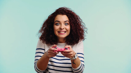 KYIV, UKRAINE - JUNE 30, 2021: young happy woman holding pink joystick and playing video game isolated on blue © LIGHTFIELD STUDIOS