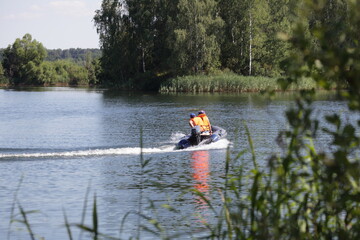 Fototapeta na wymiar Moving away inflatable motor boat with outboard motor and man in orange lifejacket fast floating on the water on green trees background at Sunny summer day, beautiful European river landscape