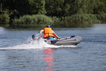 Fototapeta premium One man in orange lifejacket fast floats on inflatable motor boat with small outboard motor on the river on green grass background on shore, active recreation on the water at Sunnny summer day