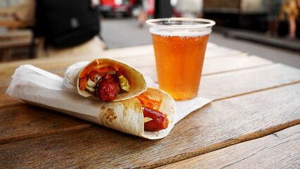 Hot dog in pita bread and a plastic glass with cider on a wooden table. Street food at the city...