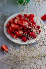 White plate with sliced strawberries and raspberries on a gray concrete background surrounded by small white flowers. Gentle spring mood, light snack
