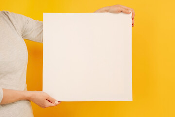 Woman holding white canvas paper blank with copyspace in her hands on yellow background. Empty sheet banner with place for text