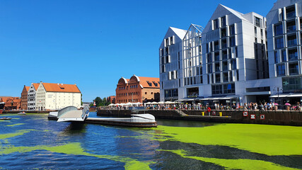 Gdansk, Poland - July 11, 2021: View of the old city of Gdansk on the Motlawa River. Tourists walk along the waterfront. On Motlawa green patches of duckweed. Middle of the river flows ship.