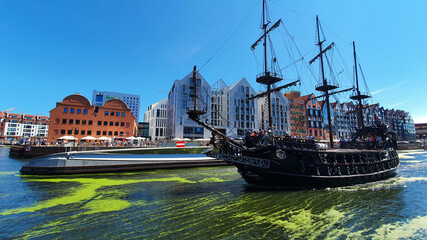 Obraz premium Gdansk, Poland - July 11, 2021: View of the old city of Gdansk on the Motlawa River. Tourists walk along the waterfront. On Motlawa green patches of duckweed. Middle of the river flows ship.