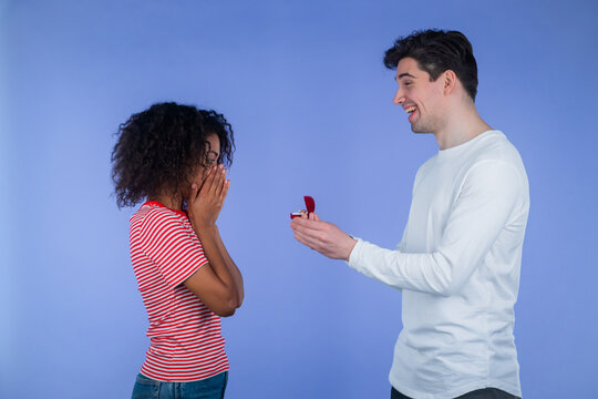 Young Interracial Couple. Man Makes Marriage Proposal To His Lover African Woman With Ring On Blue Studio Background. Love, Relationship, Happiness Concept.