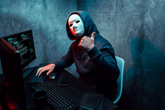 Anonymous hacker wearing face mask working on computer in dark room